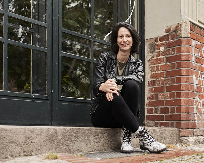 Frau Anfang 30 sitzt im Schneidersitz an Backsteinwand; trägt Lederjacke, grünes Shirt, schwarze Leggings und helle Schnürstiefel.