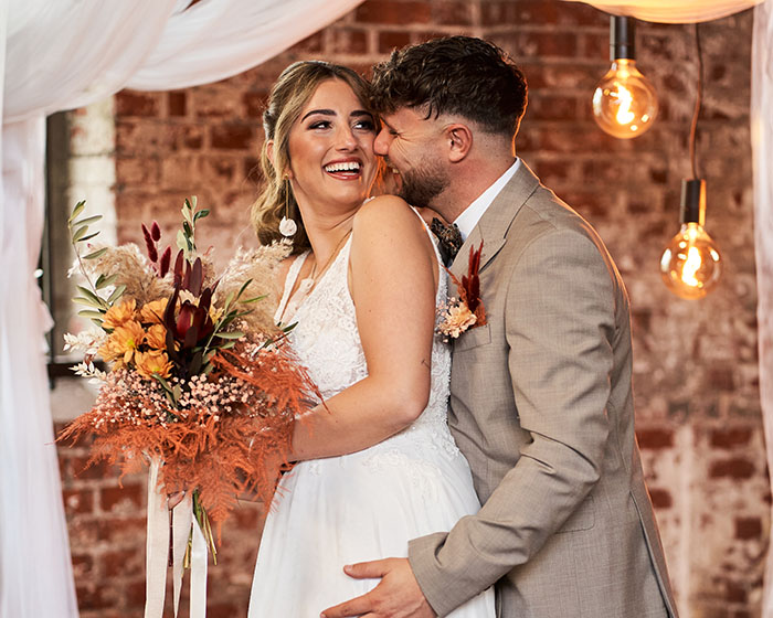 Fröhliches Brautpaar steht eng umschlungen vor Backsteinwand; sie lacht mit Blumenstrauß in der Hand, er küsst ihre Wange.