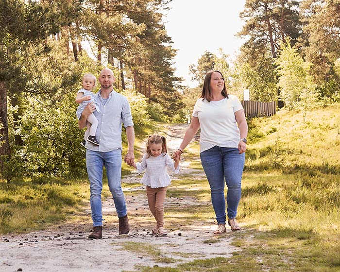 Junge Familie mit zwei kleinen Kindern geht fröhlich einen sonnigen Waldweg entlang; Eltern halten Tochter an den Händen, Vater trägt Kleinkind.