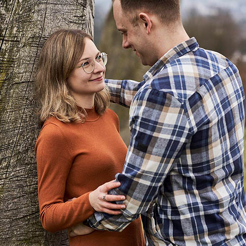 Junges Paar steht eng beieinander an einem Baum; sie lächelt ihn an, er hält sie liebevoll an der Taille.