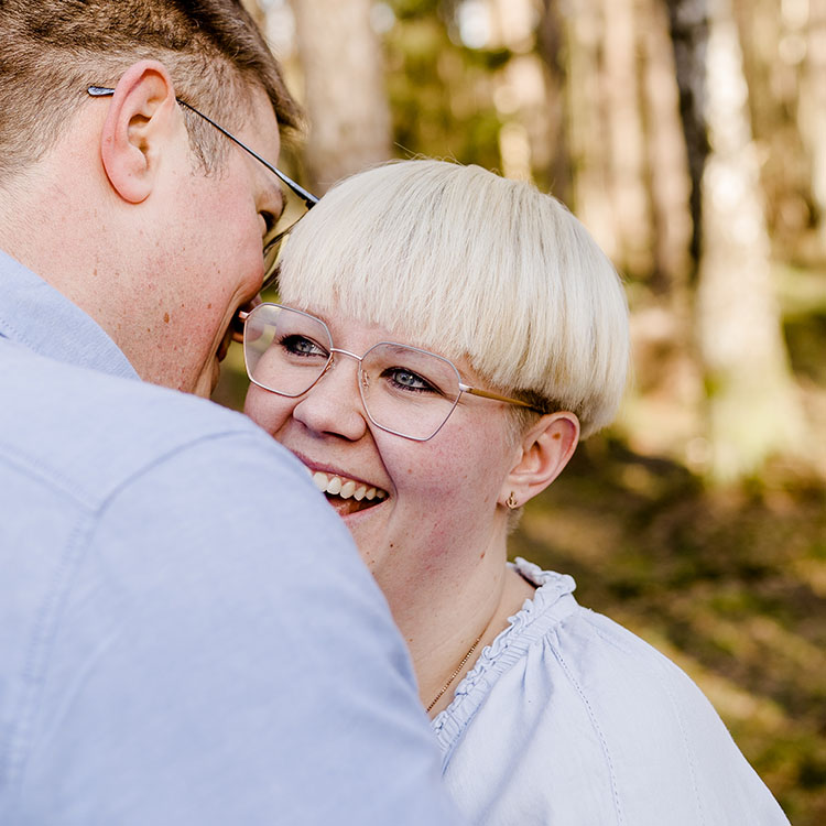 Lachende Frau mit hellem Kurzhaarschnitt und Brille blickt ihren Partner liebevoll an; vertrauter Moment im Wald.