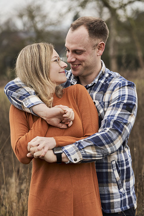 Mann umarmt Frau von hinten, beide blicken sich lächelnd in die Augen; innige Geste in herbstlicher Natur.