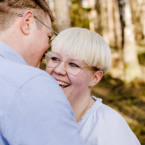 Lachendes Paar in heller Kleidung steht nah beieinander im Wald; Frau mit Kurzhaarschnitt blickt den Mann verliebt an.