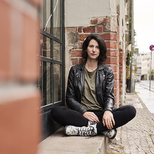 Frau Anfang 30 sitzt entspannt im Schneidersitz vor Backsteinfassade; trägt Lederjacke, T-Shirt und Sneakers.
