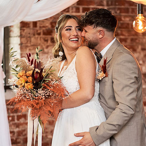 Fröhliches Brautpaar steht eng umschlungen vor Backsteinwand; sie lacht mit Blumenstrauß in der Hand, er küsst ihre Wange.