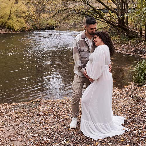Das Paar steht innig am Ufer eines ruhigen Bachs, umgeben von herbstlicher Natur. Sie lächeln sich an, seine Hände liegen behutsam auf ihrem Babybauch – ein stiller, liebevoller Moment der Vorfreude.