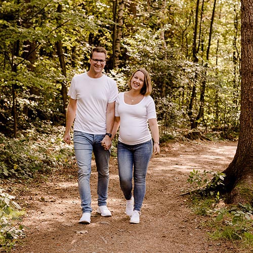 Ein Paar spaziert Hand in Hand durch einen sonnigen Waldweg. Beide tragen helle Kleidung und lächeln in die Kamera. Die Frau ist schwanger und legt eine Hand auf ihren Bauch.