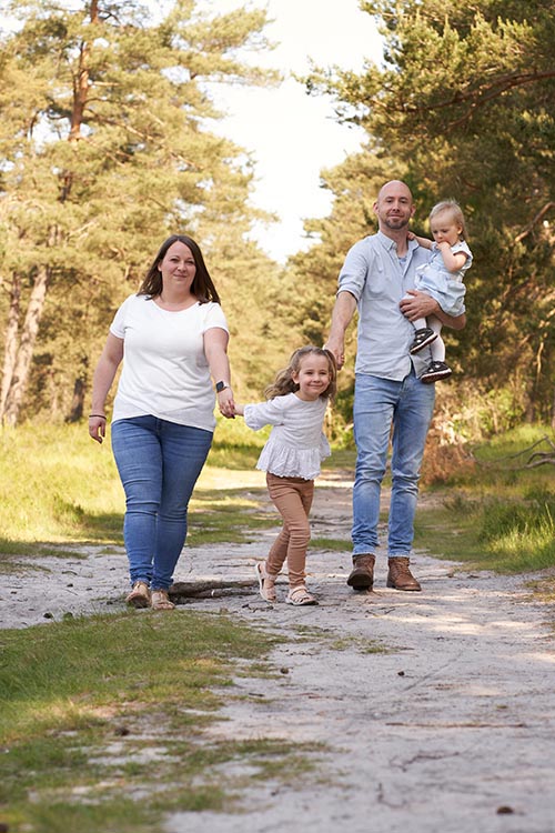Eltern gehen mit zwei kleinen Kindern durch lichten Wald; Tochter läuft zwischen den Eltern, Vater trägt das jüngere Kind.