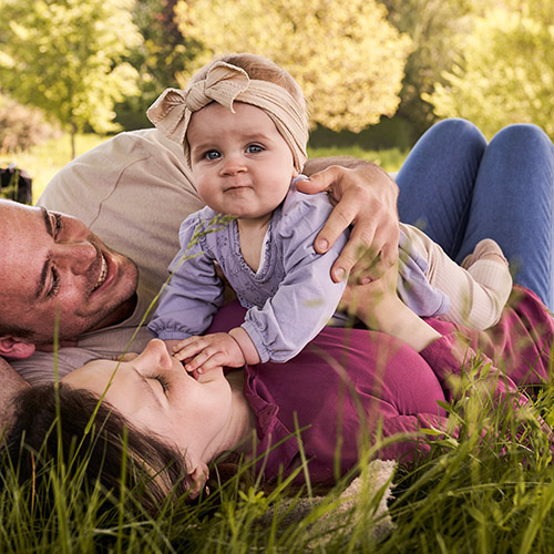 Baby mit Stirnband liegt auf Mutter, Vater daneben; alle entspannt auf Wiese, Sommerlicht und Nähe spürbar.