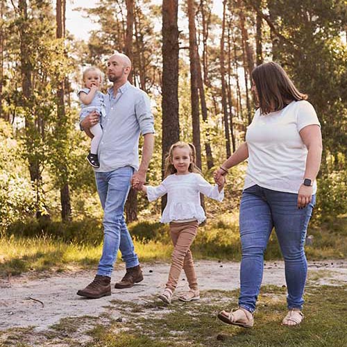 Eltern gehen mit zwei kleinen Kindern durch lichten Wald; Tochter läuft zwischen den Eltern, Vater trägt das jüngere Kind.