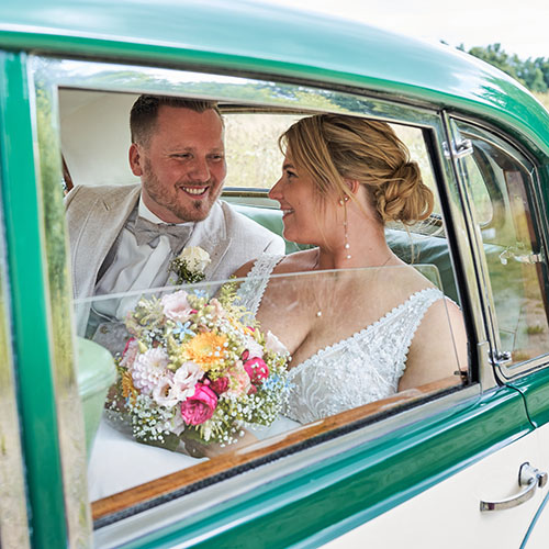 Brautpaar sitzt lächelnd im Oldtimer, schaut sich verliebt an; Braut hält bunten Strauß, beide elegant gekleidet.