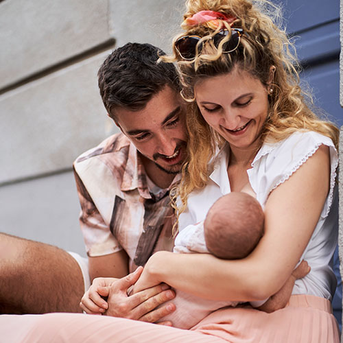 Junges Elternpaar sitzt draußen und hält ihr Neugeborenes liebevoll im Arm, beide lächeln das Baby an – inniger Moment der Familienfotografie outdoor.