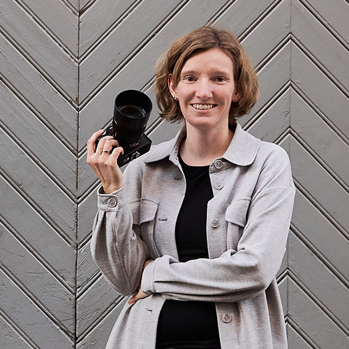 Portrait der Fotografin Annika Rose mit Kamera in der Hand vor einer grau gestrichenen Holzwand – spezialisiert auf authentische Babybauch-, Neugeborenen-, Familienfotos und Hochzeitsreportagen.