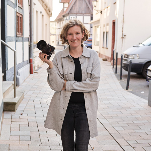 Fotografin Annika Rose lächelt freundlich in die Kamera, während sie mit ihrer Kamera entspannt durch eine Gasse in Lemgo spaziert – authentisch und herzlich.