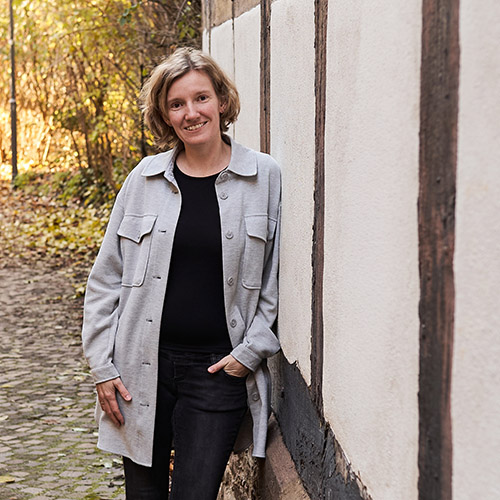 Portrait von Fotografin Annika Rose, lächelnd an eine Fachwerkhauswand gelehnt, umgeben von herbstlicher Natur in einer gepflasterten Gasse in Lemgo.
